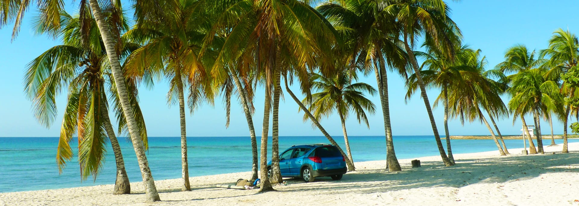 kubanische Feiertage. Landschaft Playa Chiron. Große grüne Palmen auf einem Sandstrand und Reisende im Schatten in der Nähe des blauen Autos.Playa Giron, Kuba.