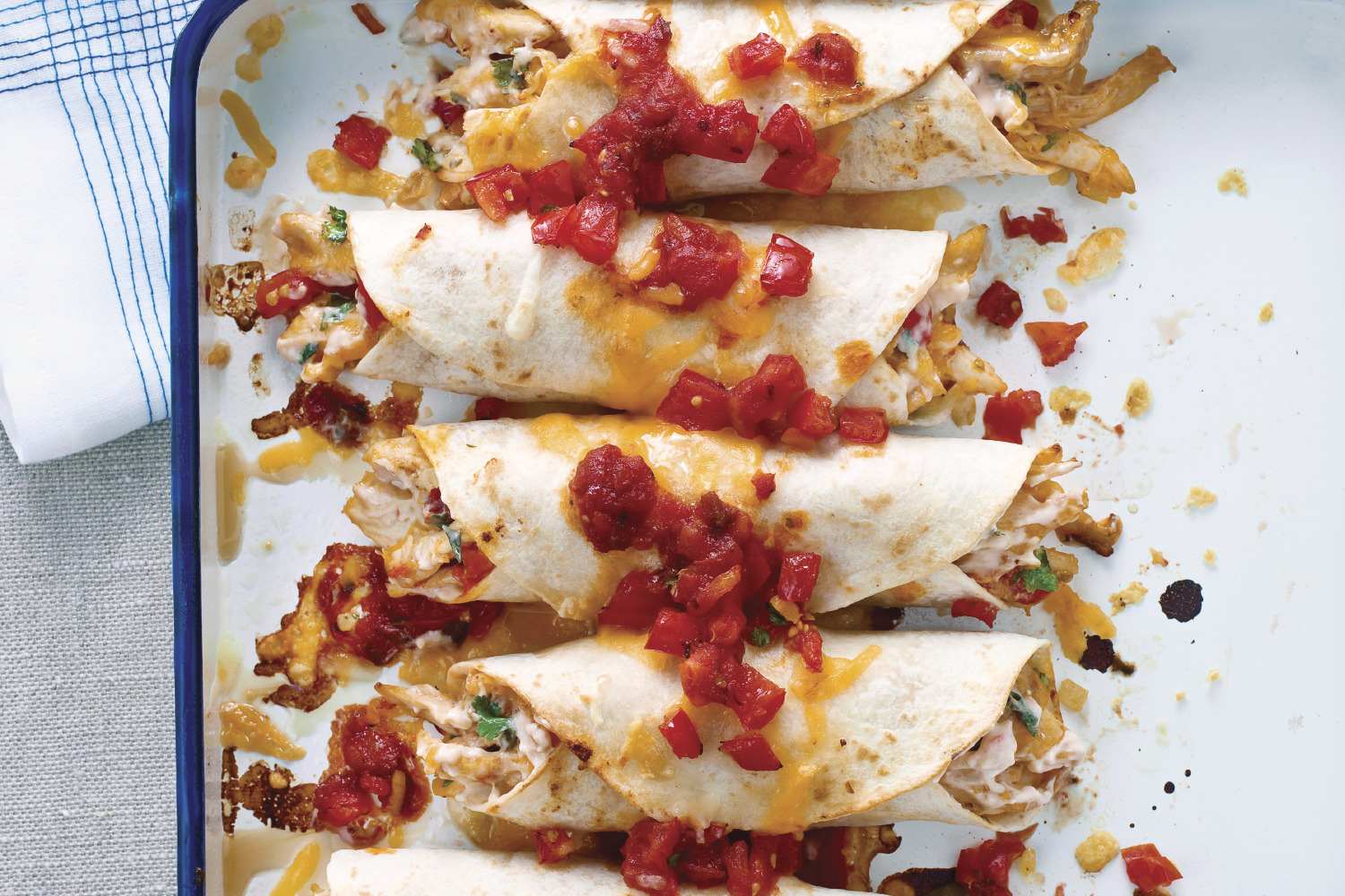 A close-up overhead shot features several chicken enchiladas arranged neatly in a white baking dish with a blue rim. The enchiladas are wrapped in lightly browned flour tortillas, topped with melted cheese, and garnished with chopped tomatoes and fresh cilantro. The dish is slightly stained with remnants of melted cheese and sauce, adding to the impression of a freshly prepared and delicious meal. A patterned napkin is partially visible in the top left corner of the frame.
