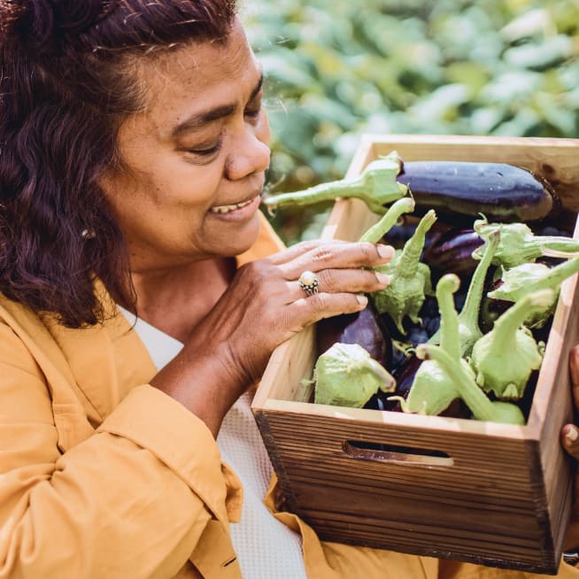 An older woman carrying a wooden box of freshly picked eggplants