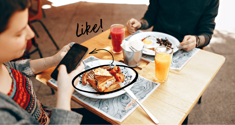 Two people sitting at a patio table outside with food and using a cellphone