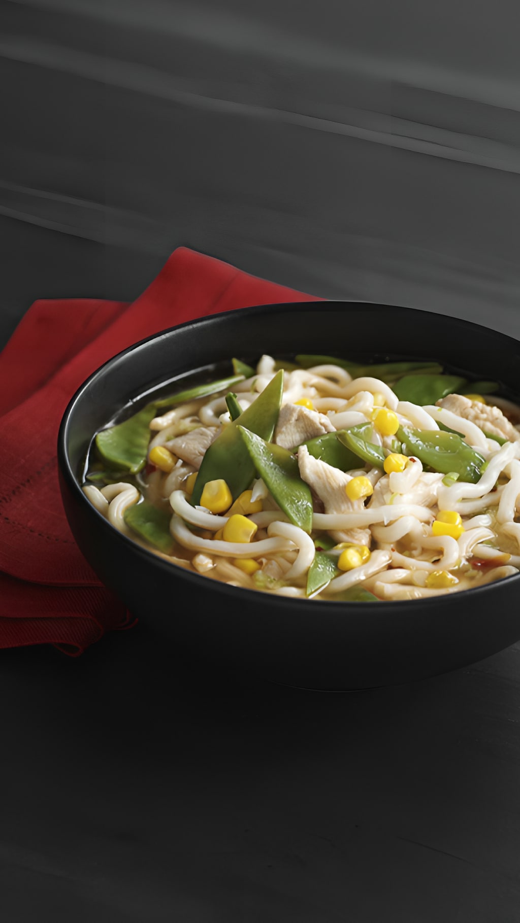 A black bowl of broth, thick noodles, snow peas, and corn, next to a red napkin.