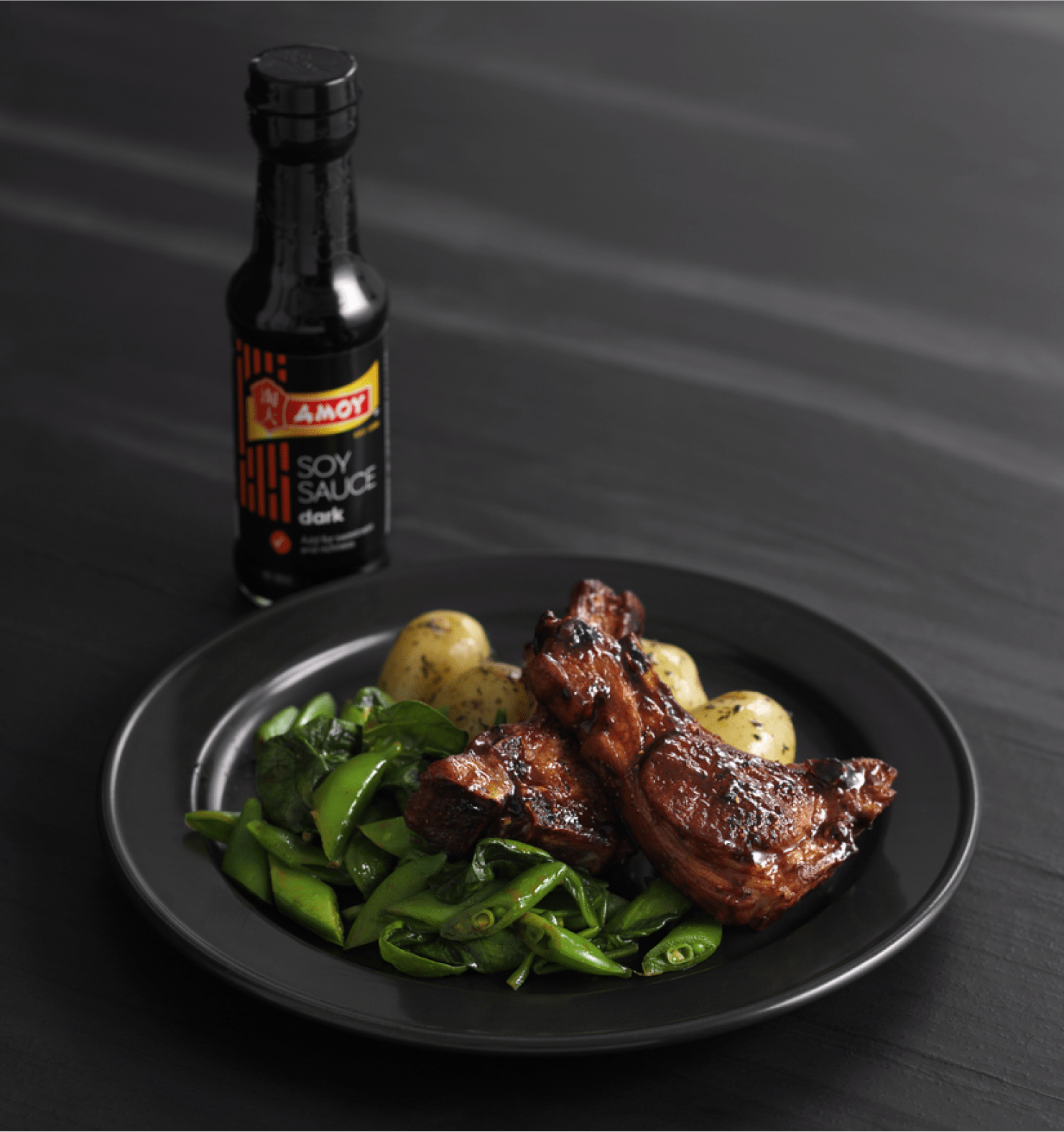 A black plate with meat, snap peas, and potatoes next to a bottle of Amoy Dark Soy Sauce.