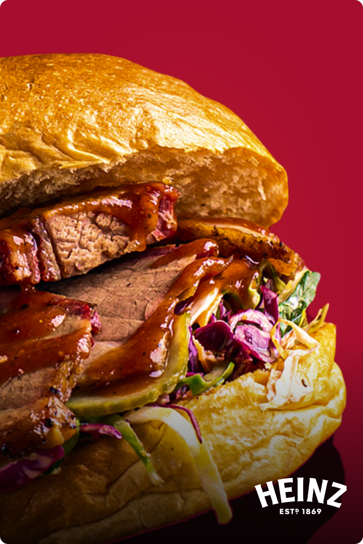 A roast beef sandwich on a crusty roll topped with pickles, cabbage, and BBQ Sauce.