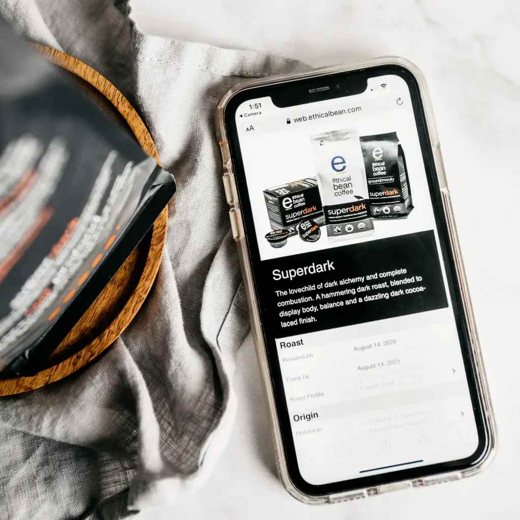 A close-up of a smartphone displaying the Ethical Bean Coffee Superdark blend product page. The screen shows images of the coffee pods and bag, along with details about the roast, origin, and flavor profile. In the background, a corner of the Superdark coffee bag rests on a wooden coaster and a soft gray cloth, further emphasizing the product. The image uses a muted color palette with black, gray, and brown tones.