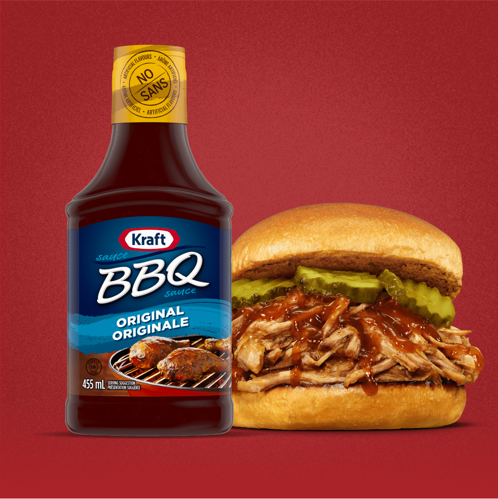 A bottle of Kraft Original BBQ Sauce is next to a pulled pork sandwich with BBQ sauce and pickles on a bun, set against a pla