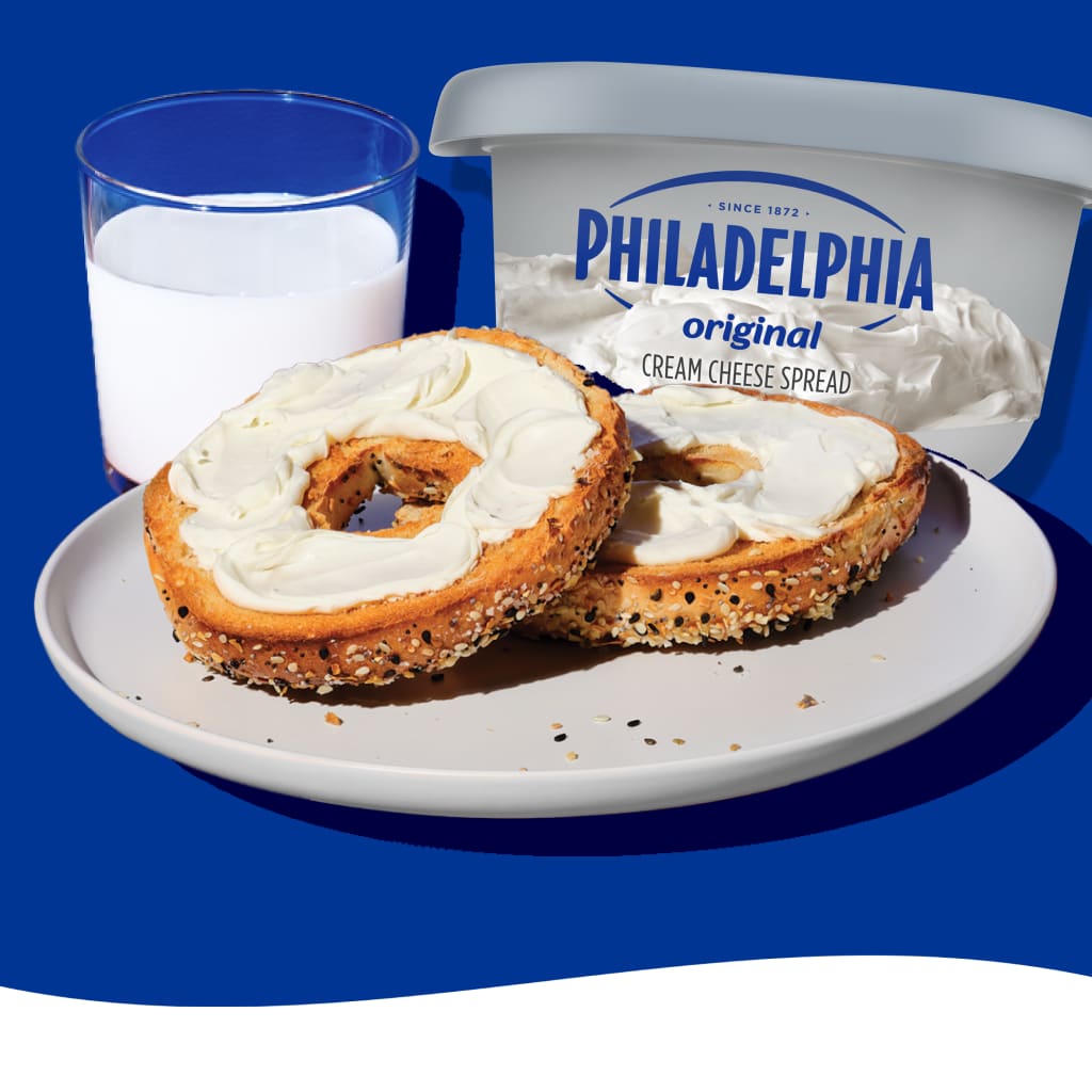 Two bagels with cream cheese, a glass of milk, and Philadelphia cream cheese.