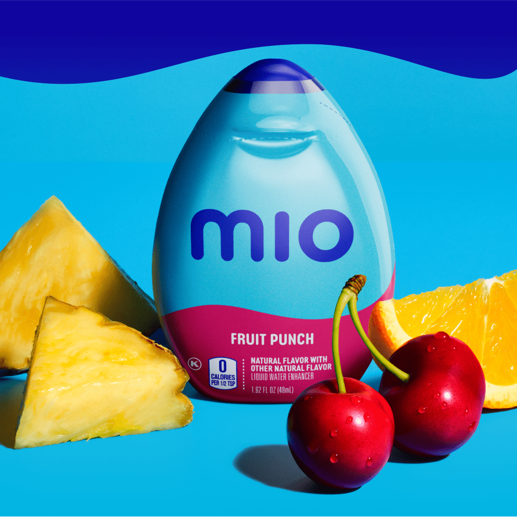 A bottle of Mio fruit punch water enhancer is placed on a blue surface, surrounded by two pineapple chunks, a slice of orange