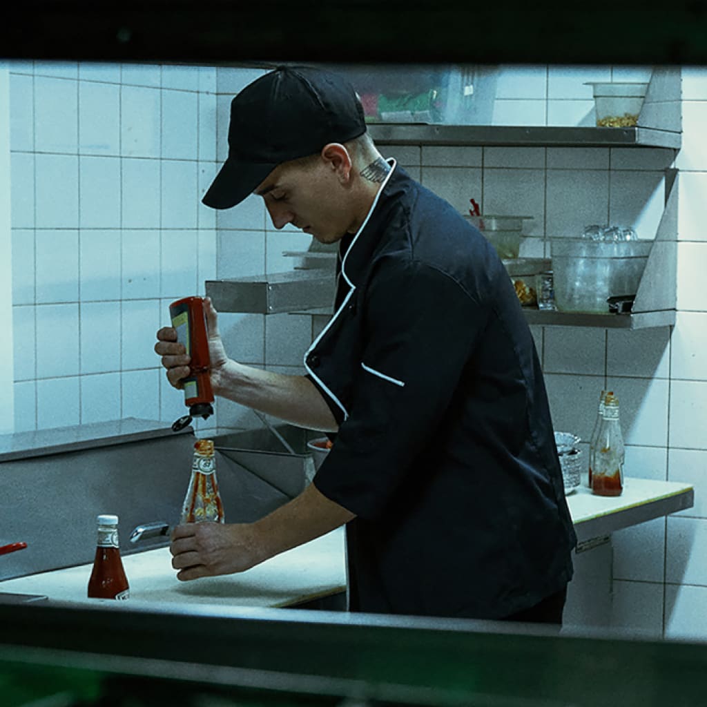 A waiter in a restaurant kitchen pours counterfeit ketchup into an empty Heinz bottle. It’s gotta be Heinz, even if it’s not.