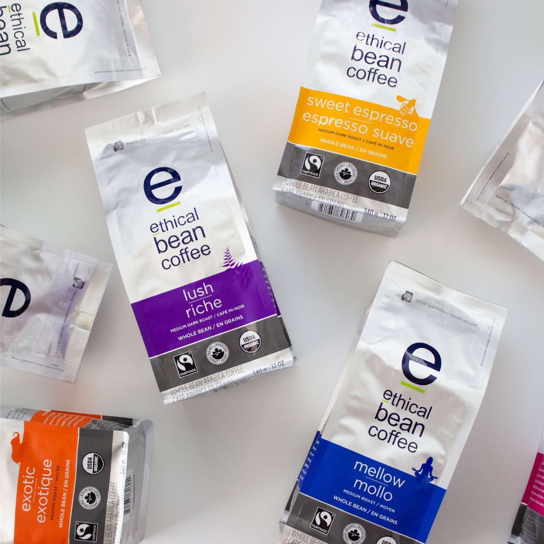 A flat lay arrangement of Ethical Bean Coffee bags in various colors, including purple, orange, gold, and blue, showcasing the different roasts and flavors available. The bags feature the Ethical Bean Coffee logo and details about each blend. The image highlights the brand's commitment to fair trade and organic practices through certifications displayed on each bag.