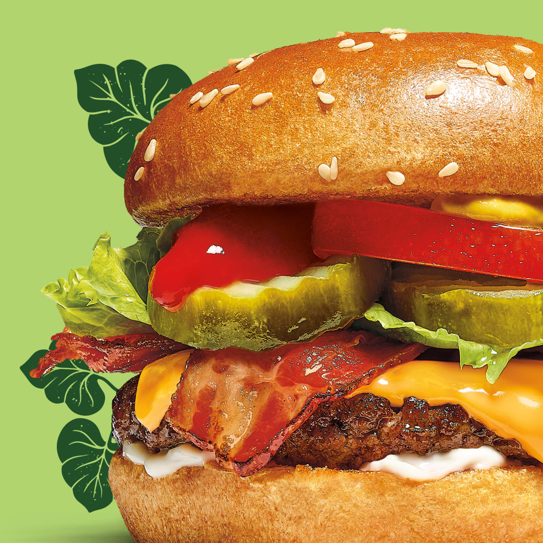 A burger with sesame bun, mustard, tomato, Claussen pickles, lettuce, bacon, cheese and a patty.