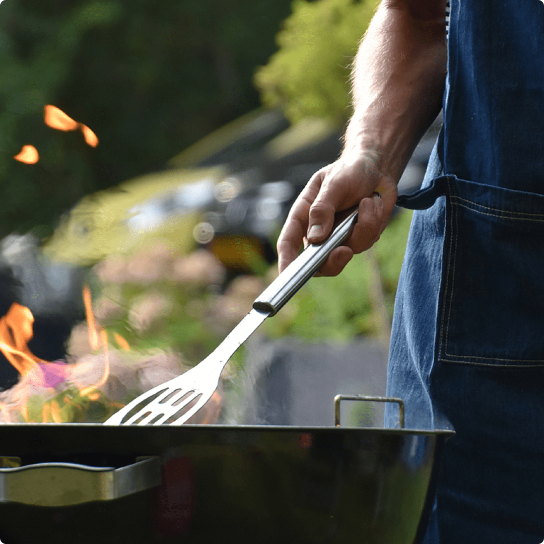 An aproned man flipping meat on a BBQ.