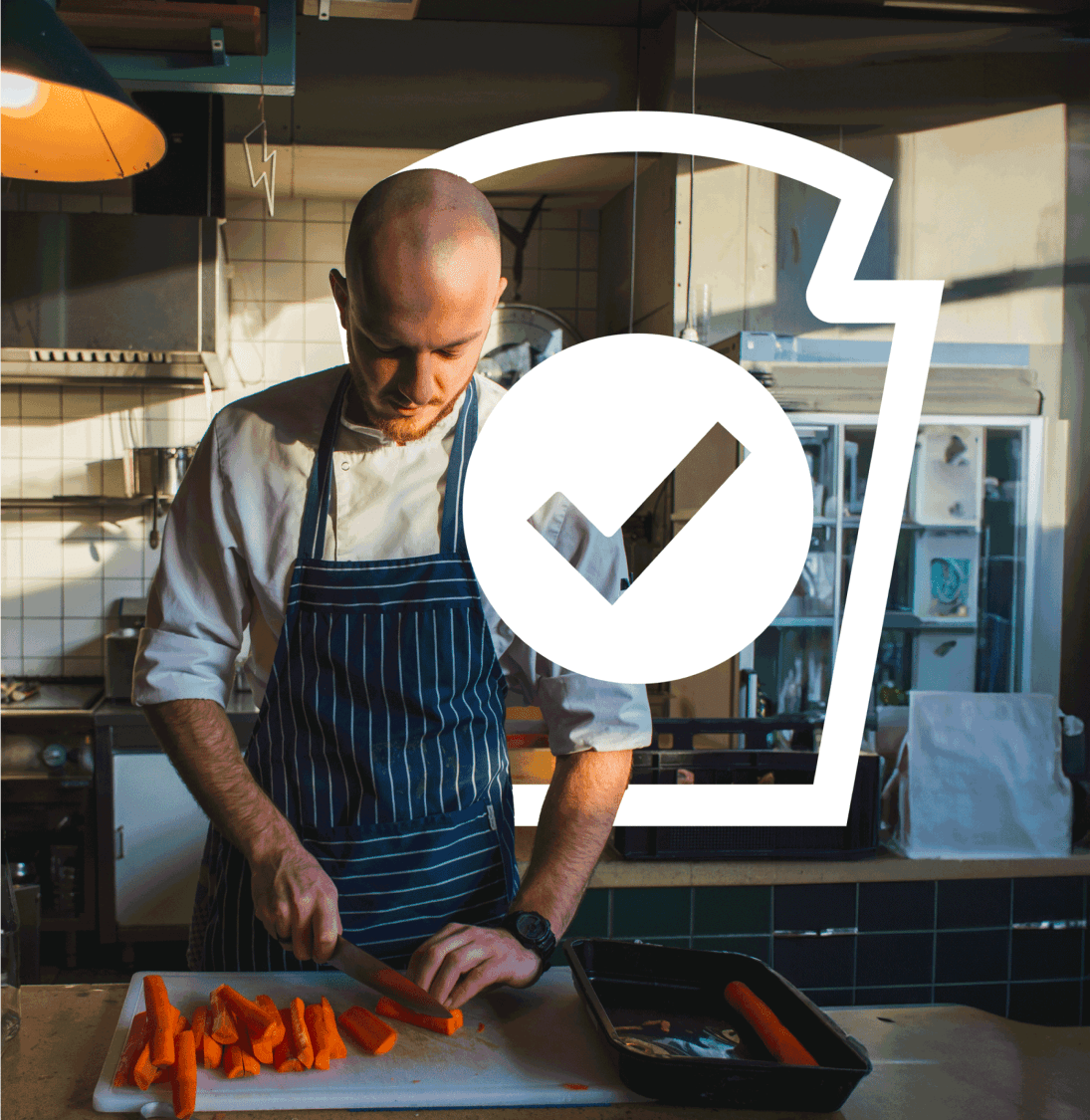 A chef wearing a striped apron carefully chops carrots on a white cutting board in a commercial kitchen, with a ticked cup gr