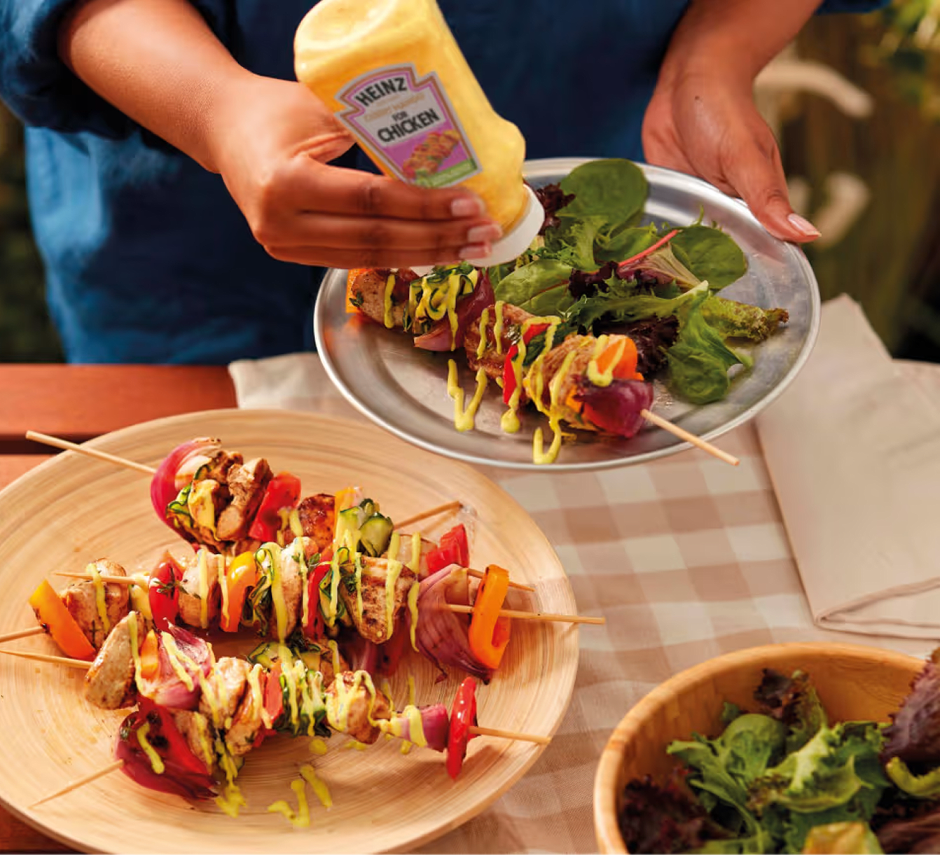 Iemand die Heinz mosterd voor kip op gegrilde kipspiesjes spuit met rode ui, paprika, courgette, salade en krielaardappelen.