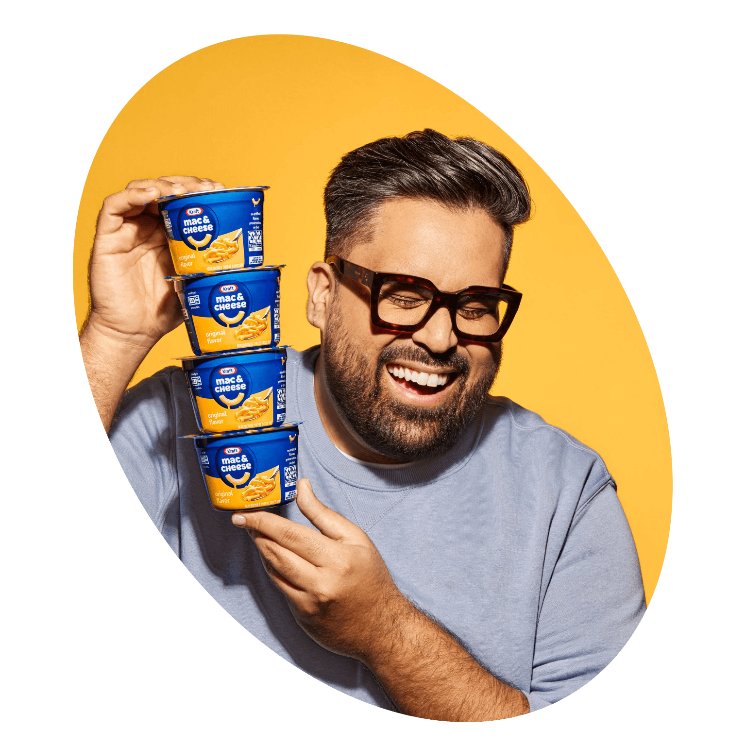 A young man with glasses smiling with his eyes closed, and holding four Kraft Mac and Cheese Cups, stacked on top of one another.