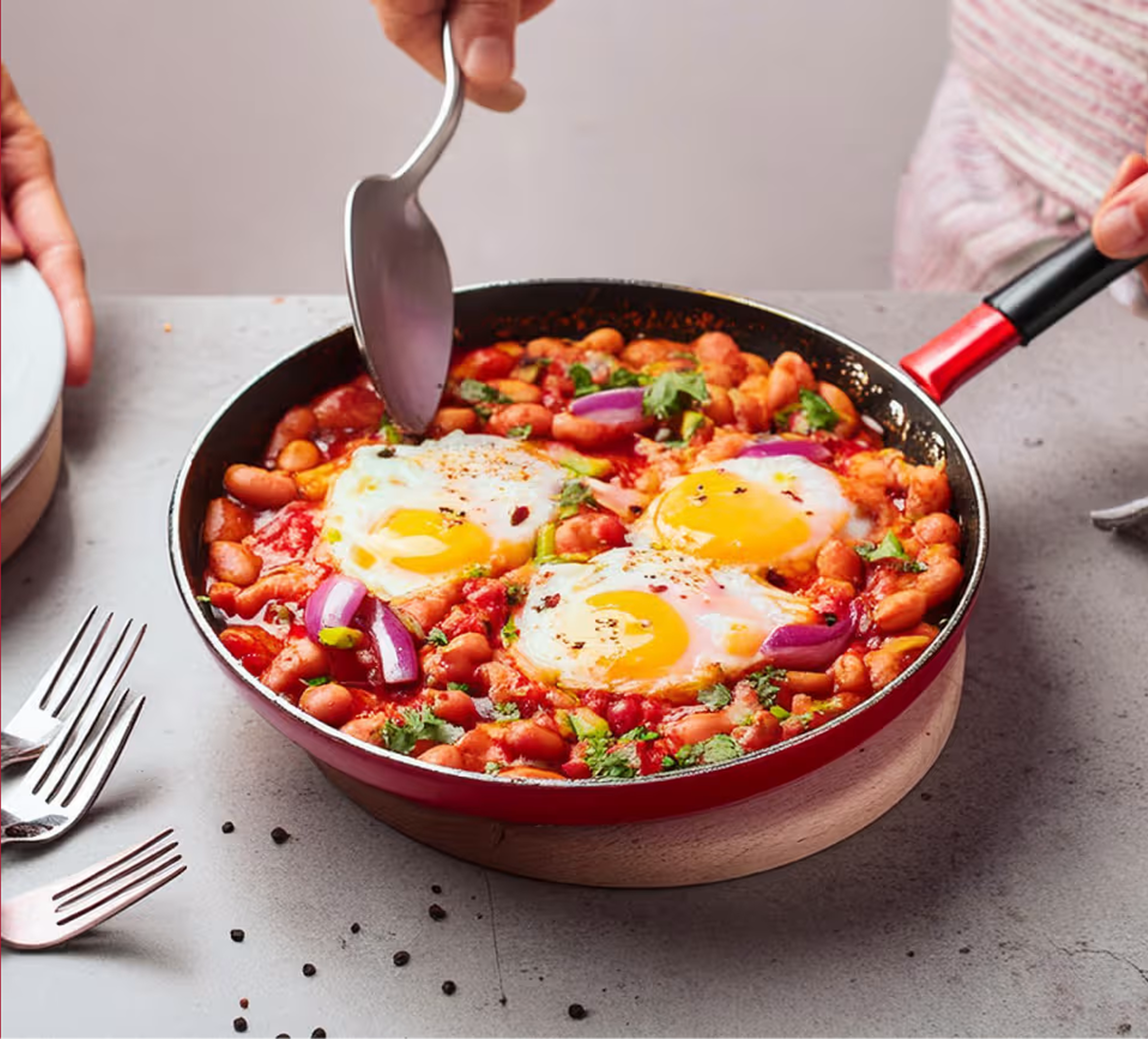 Henkilö nostaa annoksen shakshukaa punaisella pannulla. Pannu on harmaalla pöytätasolla. Tausta on punainen.