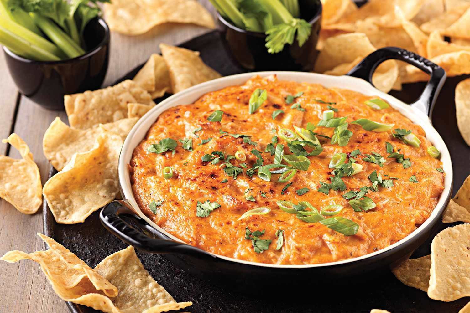 A tempting close-up of a BBQ Chicken Dip. The dip, a vibrant orange-red color, is garnished with chopped green onions and fresh cilantro. It sits in a black and white serving dish, surrounded by crispy tortilla chips and accompanied by a small bowl of celery sticks.