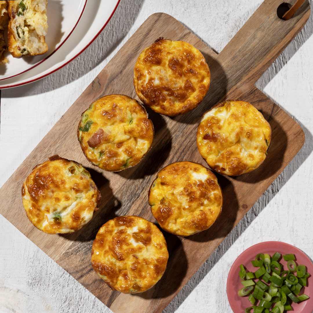A high-angle, close-up shot showcases six golden-brown, crispy Ore-Ida Tater Tot egg bites arranged on a rectangular wooden cutting board. The egg bites are nestled together, their cheesy, bubbly tops glistening under the light. Visible ingredients within the egg bites include chopped green onions and pieces of bacon.