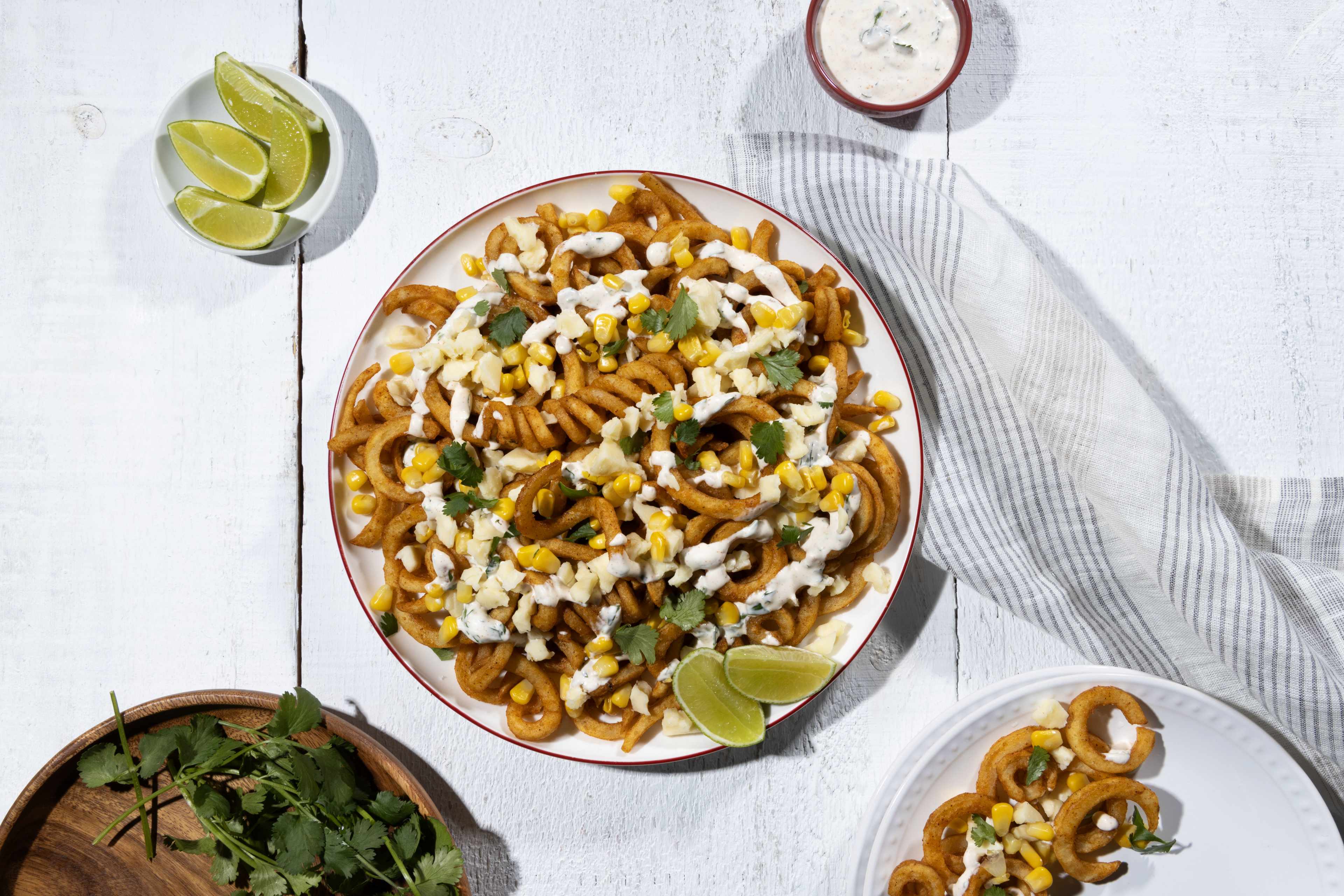 Overhead shot of a plate of seasoned curly fries topped with kernels of corn, crumbled cheese, chopped cilantro, and a creamy white sauce. Lime wedges garnish the fries, and additional lime wedges are visible in a small white bowl to the upper left. A small reddish-brown bowl of the white sauce is also present at the top, next to a folded grayish-white striped cloth napkin. A wooden bowl with fresh cilantro sprigs sits to the lower left of the main plate. A second, smaller white plate with a few curly fries and toppings is at the bottom right.