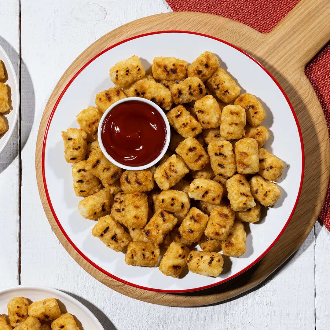 A top-down view of crispy, golden-brown Ore-Ida Tater Tots arranged on a white plate with a red rim, placed on a round wooden serving board. The tater tots have grill marks, indicating they've been cooked on a grill. A small white bowl filled with ketchup sits in the center of the plate, ready for dipping