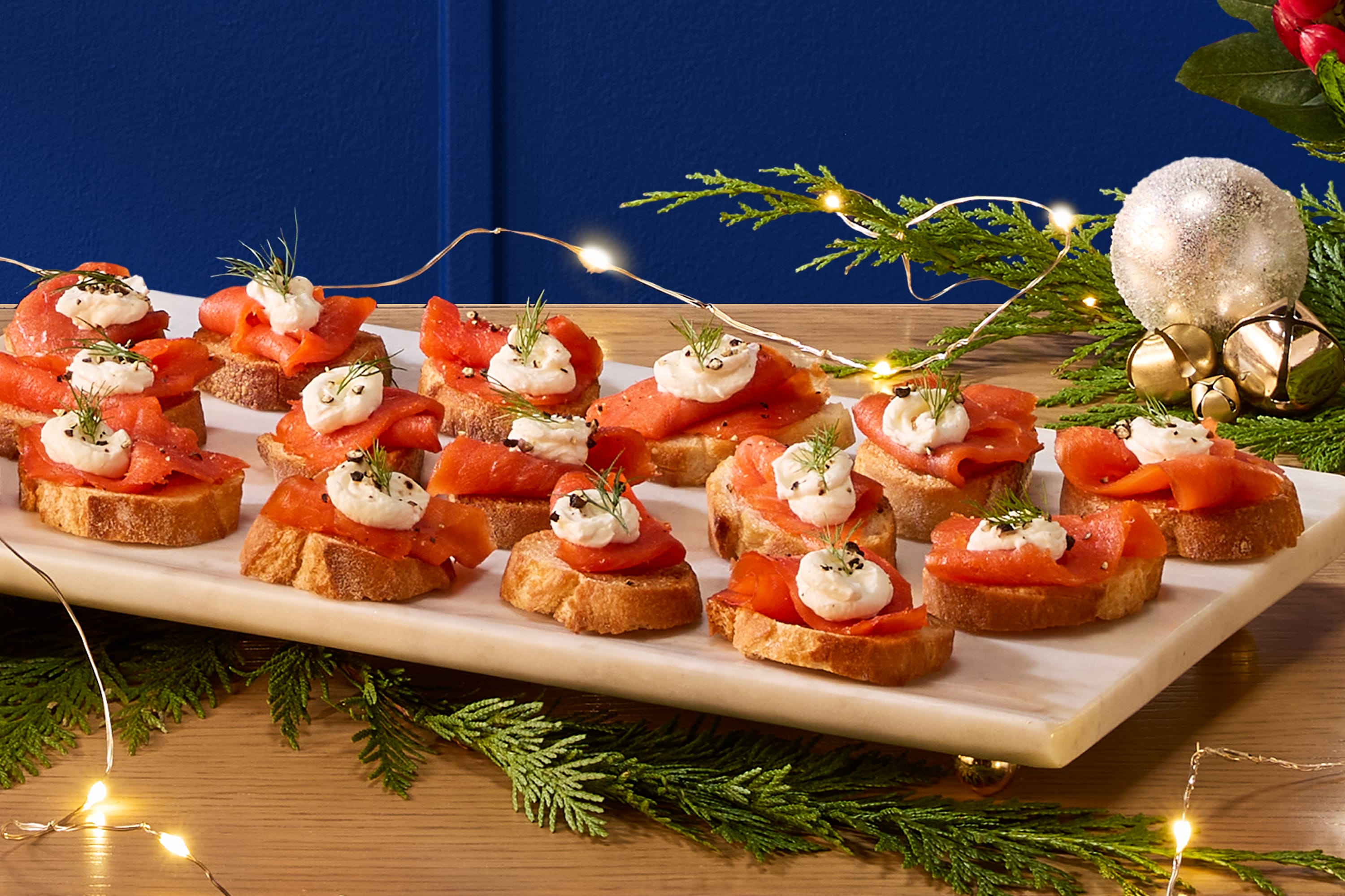 A platter of elegant Philadelphia Smoked Salmon Canapés, featuring toasted baguette slices topped with savory smoked salmon, a dollop of creamy Philadelphia cream cheese, and a fresh sprig of dill, set amidst festive holiday decorations.