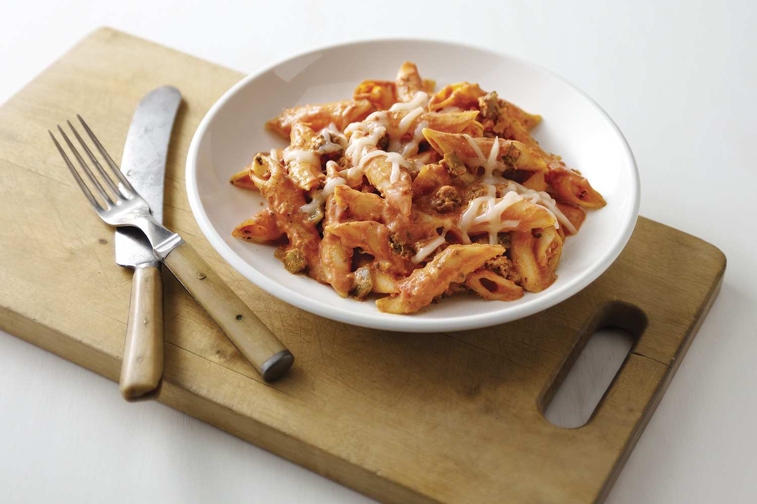 : A plate of penne pasta in a creamy tomato sauce, topped with melted mozzarella cheese. The dish is served on a white plate, which rests on a wooden cutting board. A fork and knife are placed beside the plate, ready to be used.