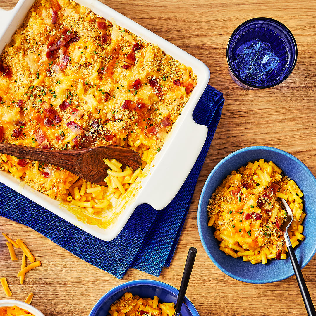 A top-down view of a rectangular white baking dish filled with creamy baked macaroni and cheese, topped with crispy bacon, golden-brown breadcrumbs, and fresh chives. A wooden spoon is resting in the dish, having served some of the mac and cheese. The casserole sits on a blue napkin on a wooden table. Two blue bowls are filled with individual servings, and a blue glass with ice water is also on the table. The scene is warm, inviting, and appetizing, showcasing a classic comfort food meal.
