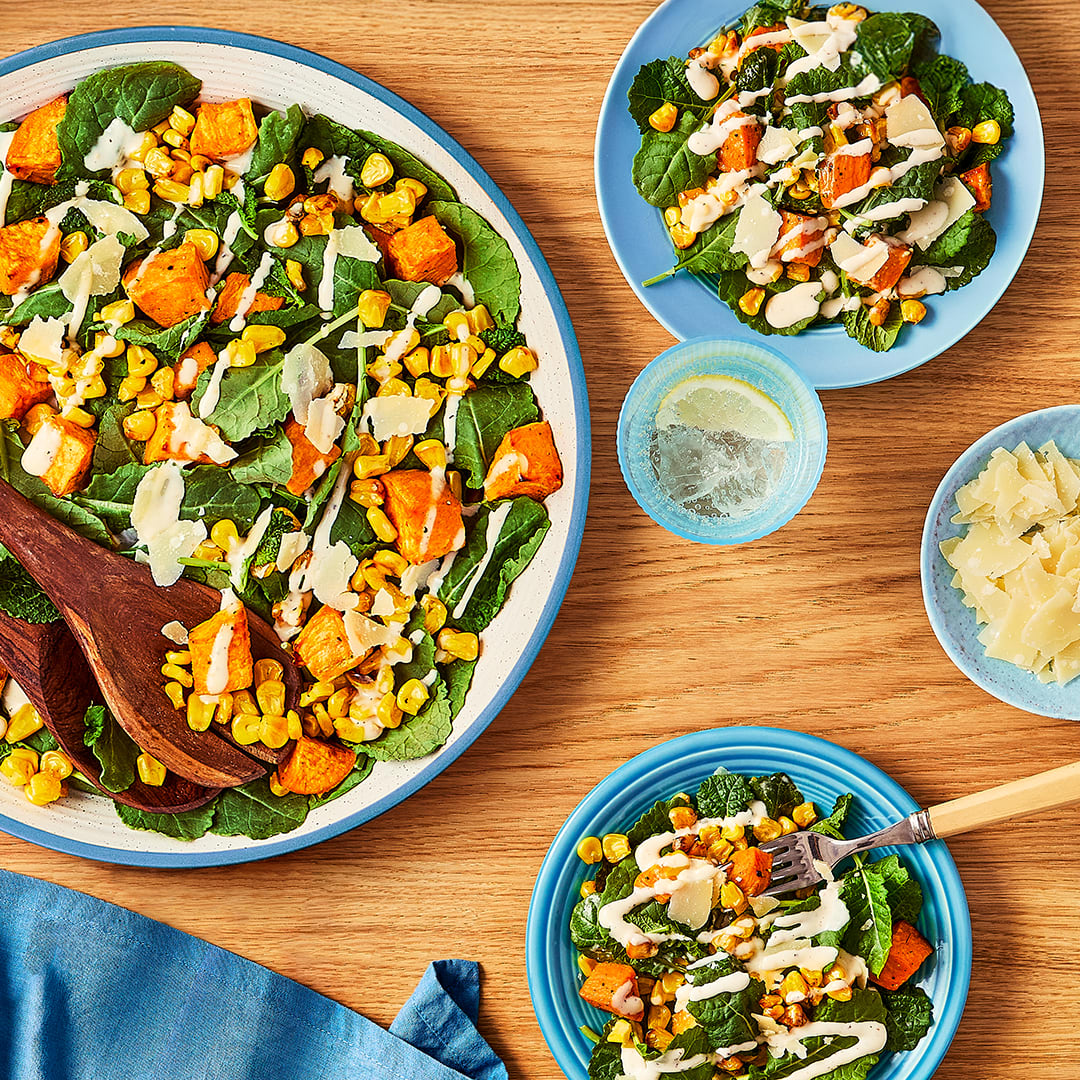 An overhead shot captures a colourful salad, featuring roasted sweet potatoes, corn kernels, and fresh kale, drizzled with a creamy dressing.