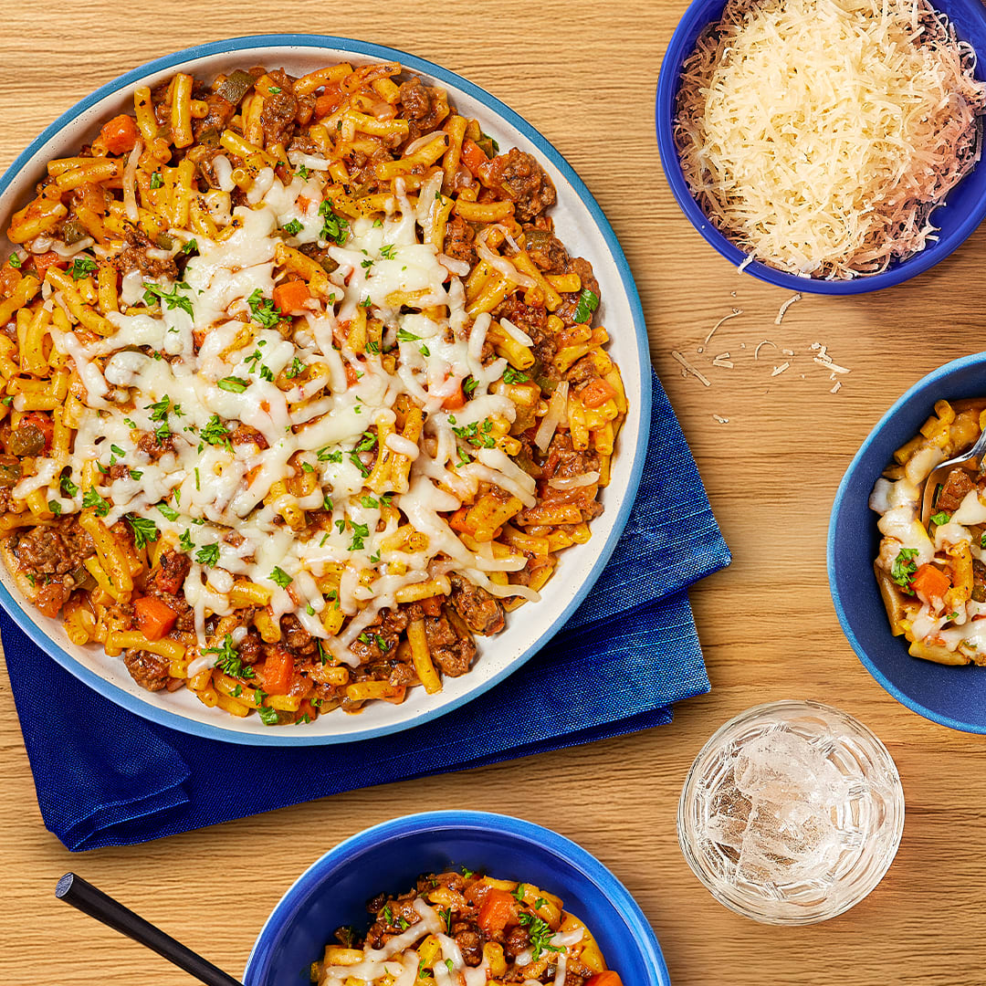 A top-down view of a delicious, cheesy one-pan bolognese macaroni and cheese dish. The main dish, served in a large bowl, is surrounded by smaller bowls containing the same cheesy macaroni, a bowl of grated parmesan cheese, and a glass of ice water.