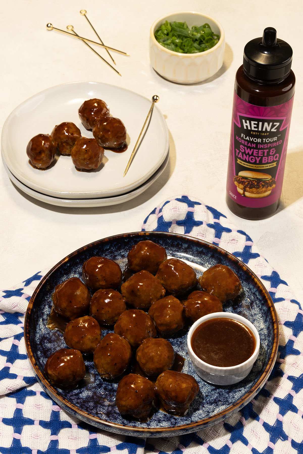 Glazed meatballs are arranged on a dark blue plate, accompanied by a small white bowl of dipping sauce. A bottle of Heinz Kor