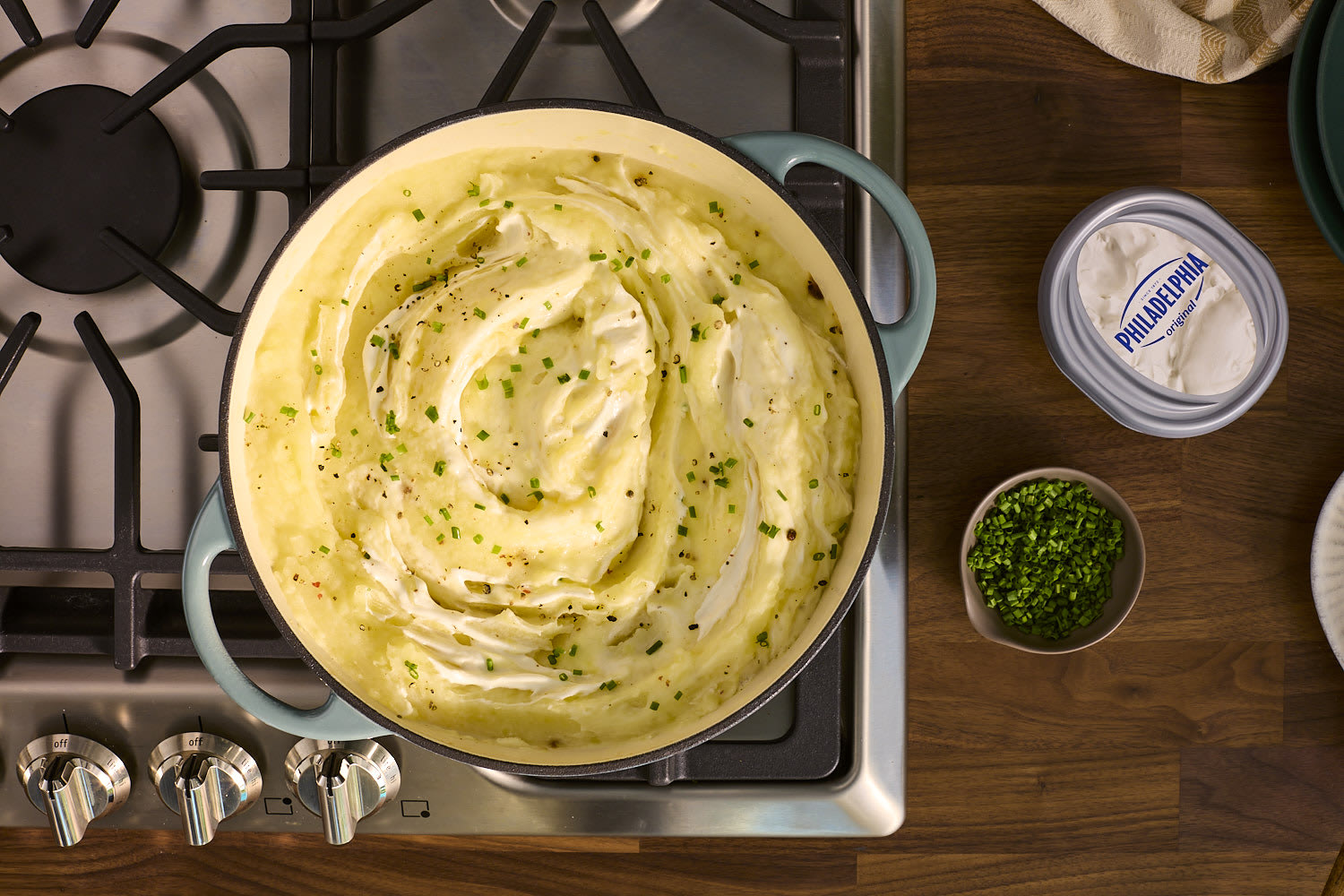 Creamy mashed potatoes topped with Philadelphia cream cheese and chives are shown in a pot on a stovetop.