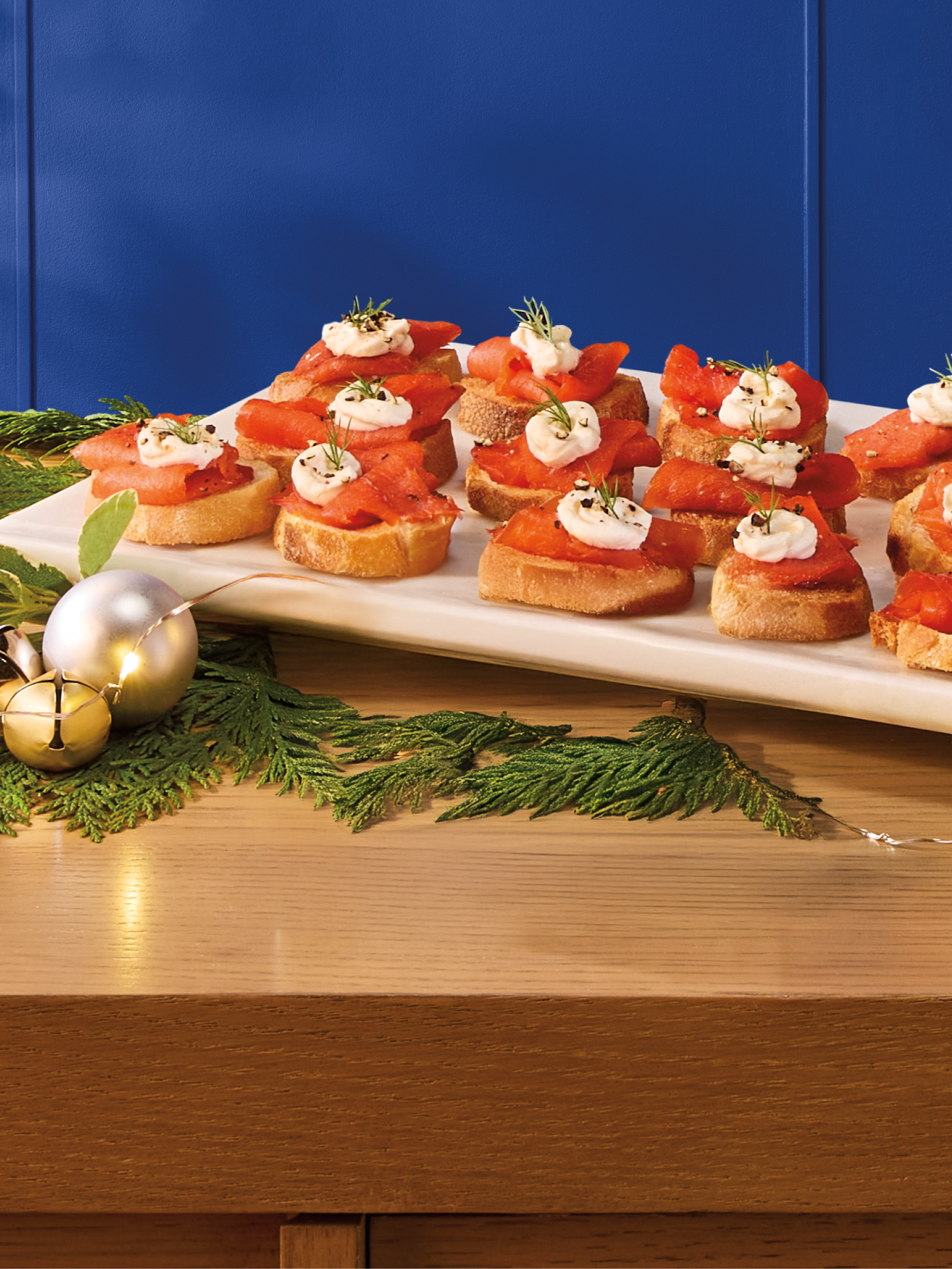 A platter of smoked salmon and cream cheese crostini garnished with pepper and dill, on a wooden table with greenery.