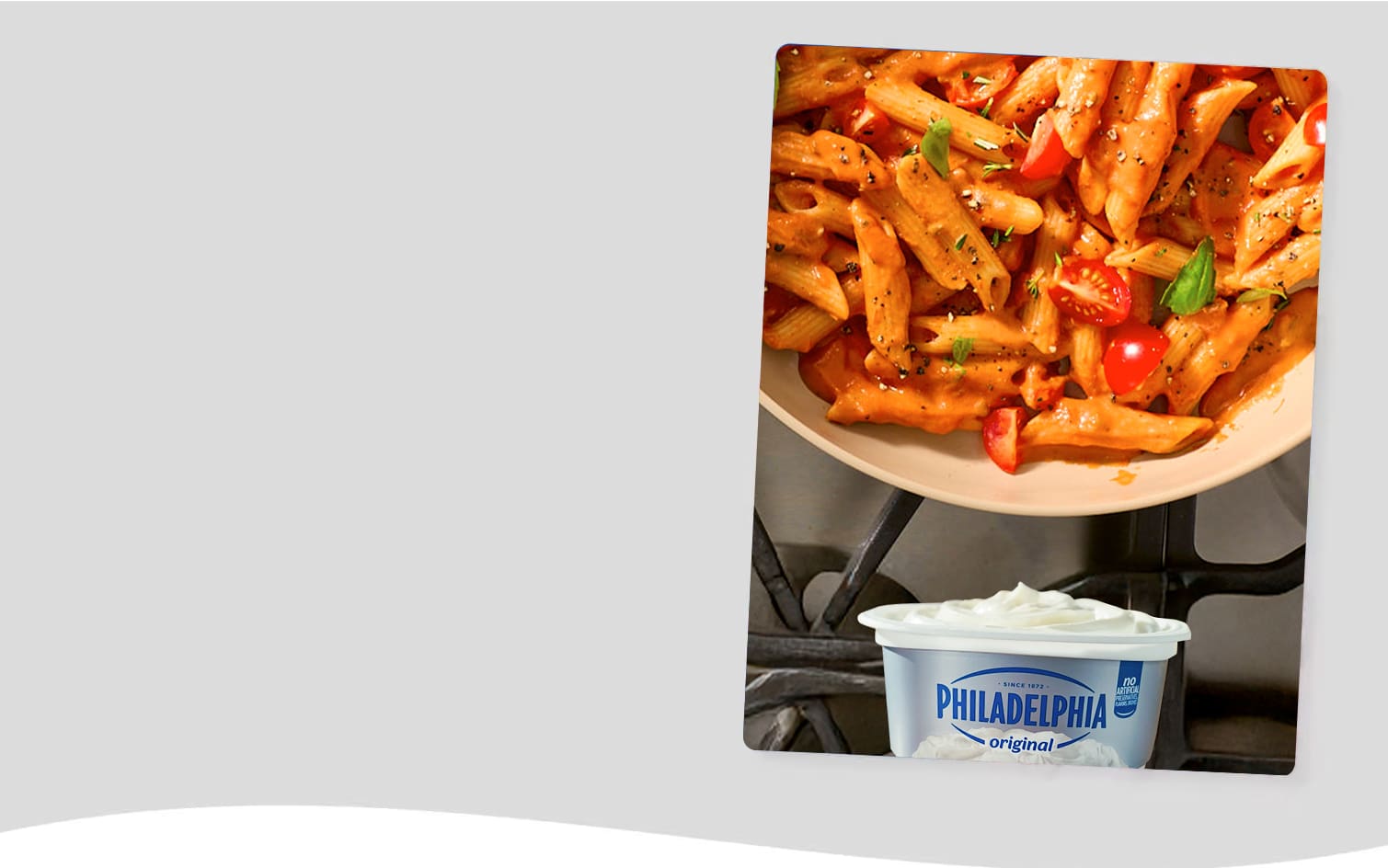Close-up of a skillet filled with penne pasta in a creamy tomato sauce, topped with fresh basil and halved cherry tomatoes. A container of Philadelphia Original Cream Cheese sits in the foreground, suggesting its use in the recipe. The warm, inviting colors of the pasta dish contrast with the clean, bright packaging of the cream cheese. The image emphasizes the creamy texture and freshness of the ingredients.