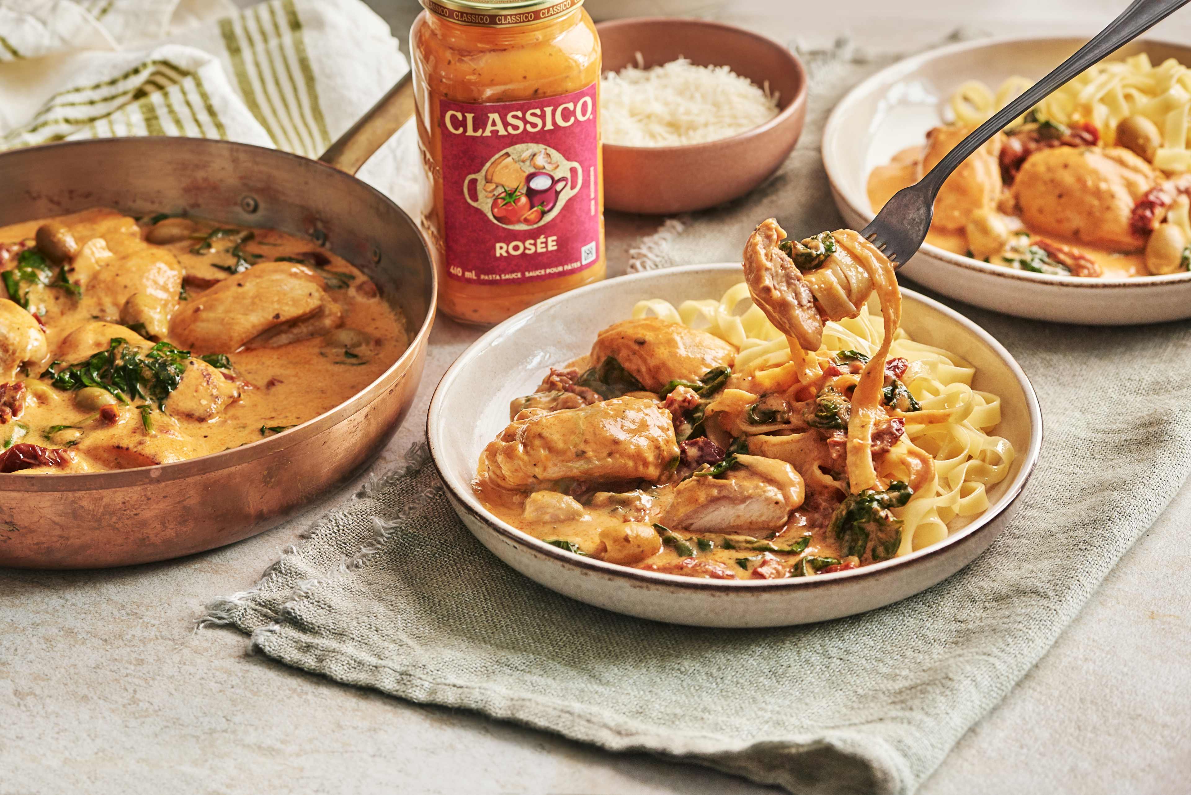 Deux assiettes de pâtes crémeuses au poulet, agrémentées de sauce rosée, d'épinards, de tomates séchées et d'olives vertes. Une fourchette enroule des pâtes et du poulet. Un pot de sauce Classico Rosé et un bol de parmesan complètent l'image.