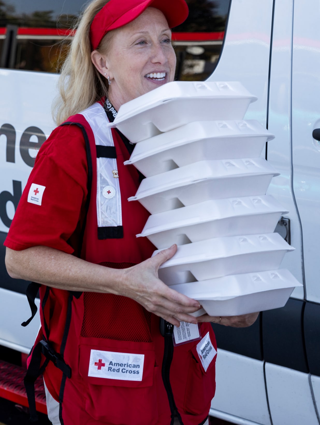 A smiling, fair-skinned woman wearing a red American Red Cross hat and vest carries a stack of white styrofoam food containers. She stands next to a white van, likely distributing meals to those in need.