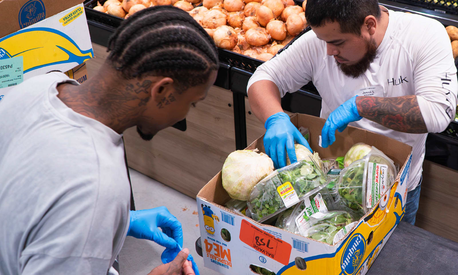 Two grocery store employees wearing blue gloves stock fresh produce. They are working with a box filled with lettuce and other leafy greens. In the background, shelves stocked with onions, sweet potatoes, and other produce are visible. The scene is bright and bustling, reflecting the activity of a busy grocery store.