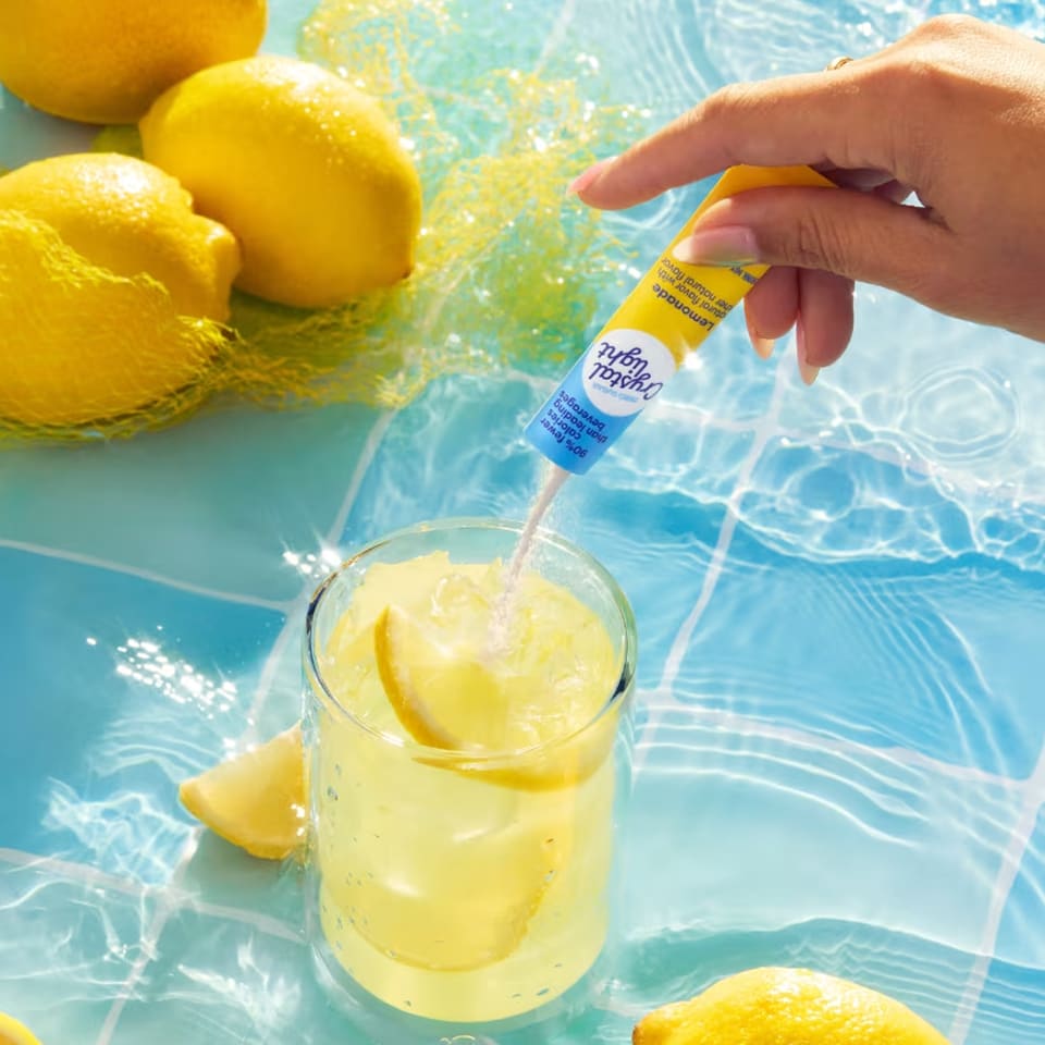 A hand pours Crystal Light Lemonade powder into a glass with ice and lemon slices