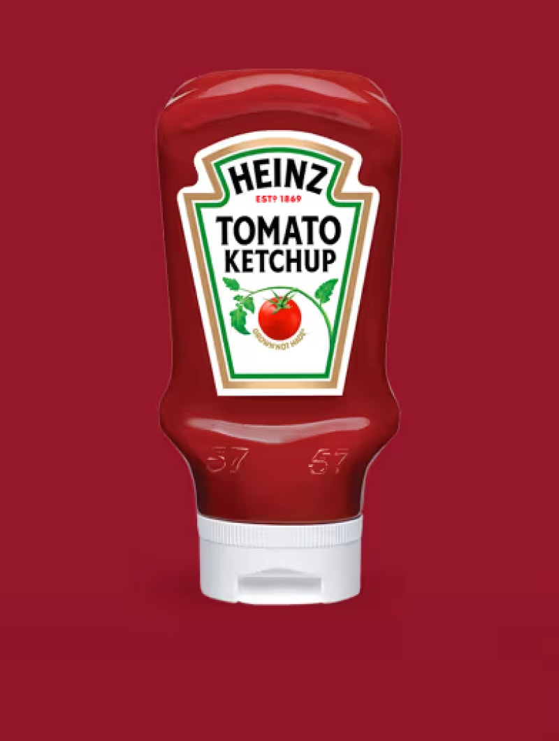 Uma garrafa squeeze de Ketchup de Tomate Heinz, vermelha, vibrante e brilhante, invertida, é exibida de forma proeminente contra um fundo vermelho escuro, mate e sólido. O rótulo clássico da Heinz, em forma de keystone, é branco com uma borda verde e dourada. 'HEINZ' está escrito em fonte sans-serif branca dentro de uma forma verde escura no topo do rótulo, com 'ESTD 1869' em pequenas letras douradas por cima. Abaixo disto, 'TOMATO KETCHUP' está em letras maiúsculas sans-serif pretas, grandes e a negrito. Uma ilustração detalhada de um tomate vermelho maduro num ramo verde com folhas encontra-se ao centro. Por baixo do tomate, 'GROWN NOT MADE' está escrito em pequeno texto branco numa faixa vermelha que acompanha a curva do tomate. A garrafa apresenta o icónico '57' em relevo nos ombros e é coberta com uma tampa flip-top branca, texturizada e limpa