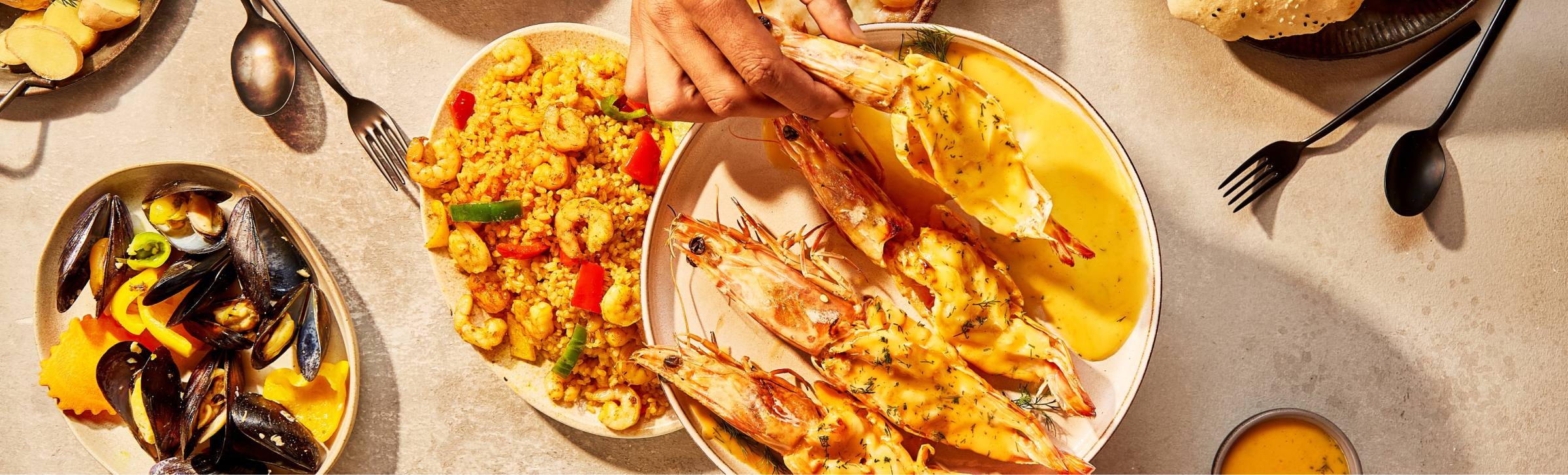 Overhead view of a table with various seafood dishes, including grilled shrimp, bulgur, bread, hummus, and drinks.