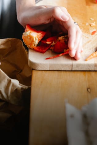 A hand holding discarded red bell pepper scraps over a wooden cutting board, with a paper bag nearby.