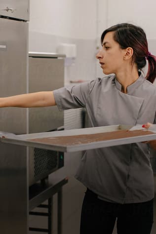A baker is seen holding a tray with what appears to be a thin sheet of chocolate or pastry, before placing it in an oven.