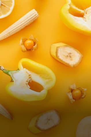 Flat lay of yellow fruits and vegetables, including lemon, baby corn, golden berries, banana slices, and bell peppers, agains