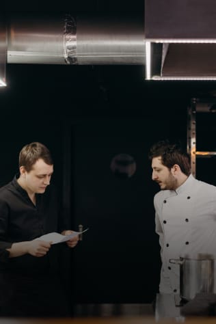 Two men in a professional kitchen. One dressed in a black shirt reading a document, while the other wears a chef's uniform.