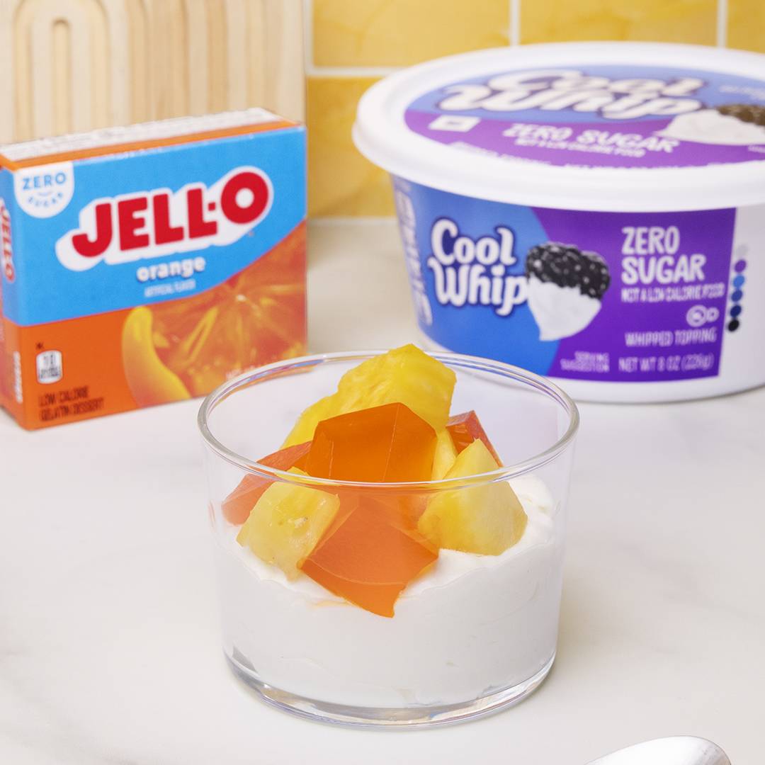 A clear glass cup filled with a creamy white base topped with vibrant orange Jell-O cubes and bright yellow pineapple chunks. This refreshing treat is presented on a light-colored countertop. In the background, a blue box of "Jell-O orange" (Zero Sugar) and a tub of "Cool Whip Zero Sugar" whipped topping are visible.