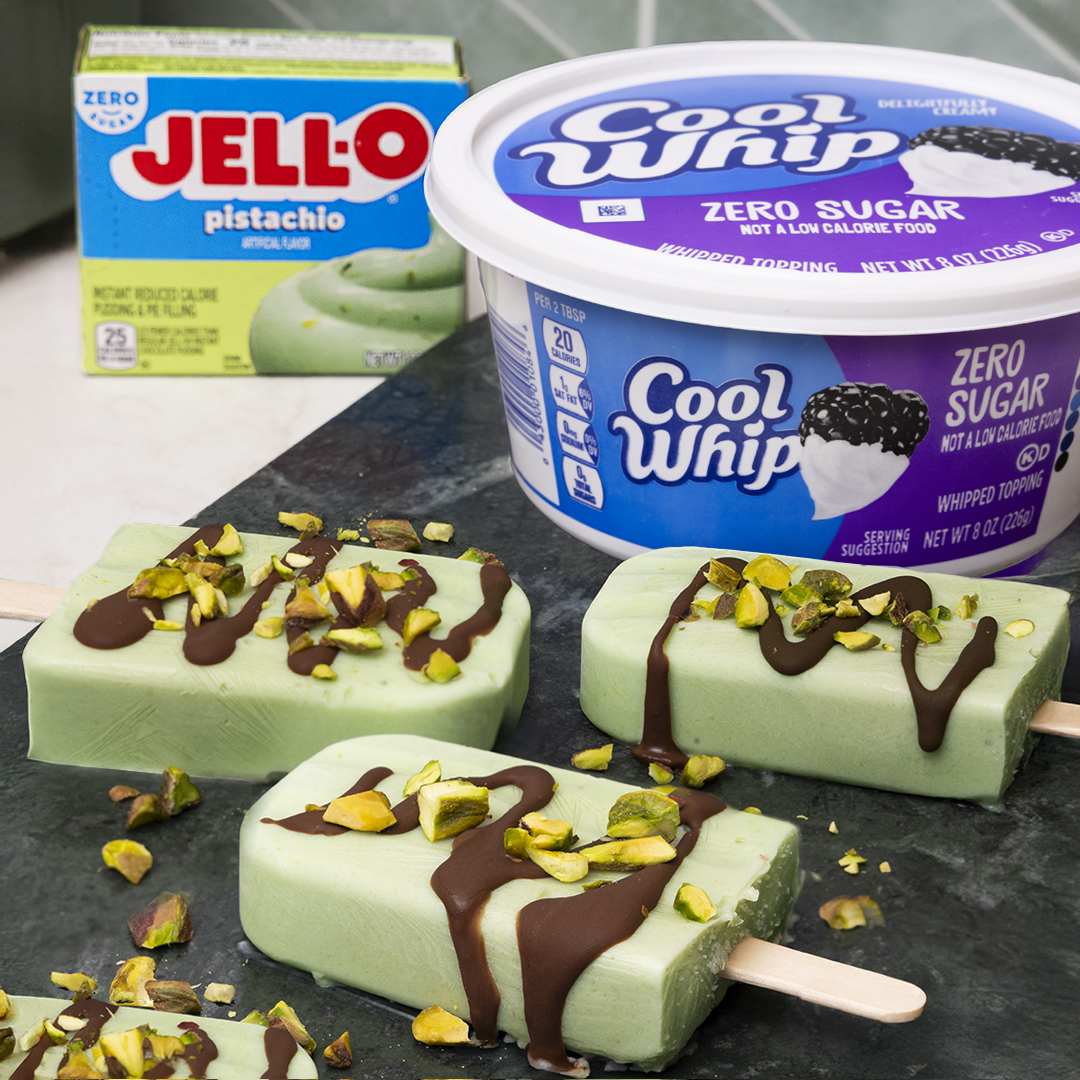 Several rectangular light green frozen pops, made with pistachio pudding, are artfully arranged on a dark, textured surface. Each pop is generously drizzled with rich dark chocolate and sprinkled with crunchy chopped pistachios. In the background, a blue box of "Jell-O Pistachio" pudding mix and a tub of "Cool Whip Zero Sugar" whipped topping are visible, hinting at the ingredients used to create these refreshing treats.
