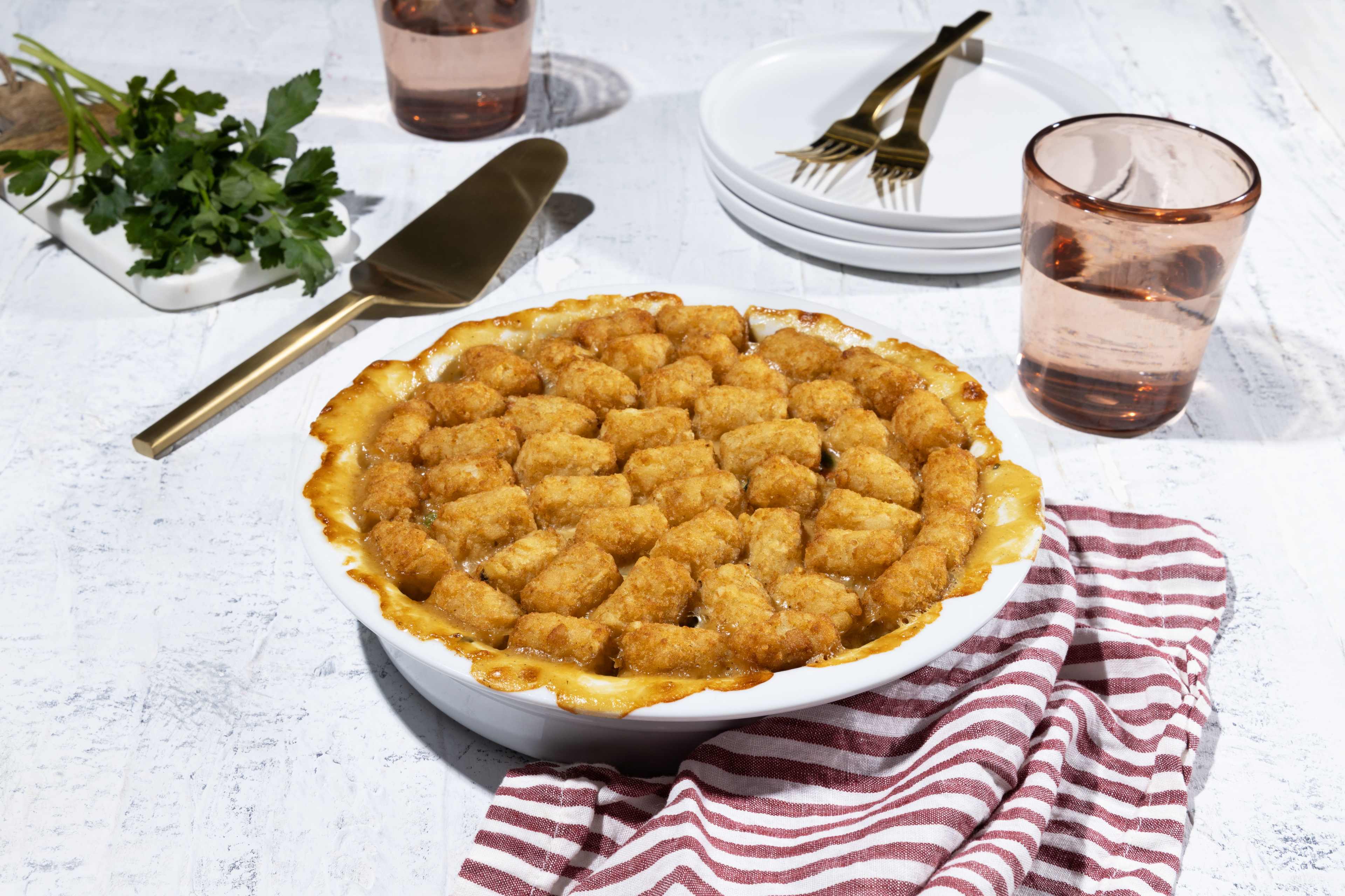 A freshly baked chicken pot pie, topped with golden-brown Ore-Ida Tater Tots, takes center stage in this inviting image. The pie, in a white baking dish, sits on a lightly textured white surface. A gold serving spatula rests beside the pie, ready to dish out a portion.