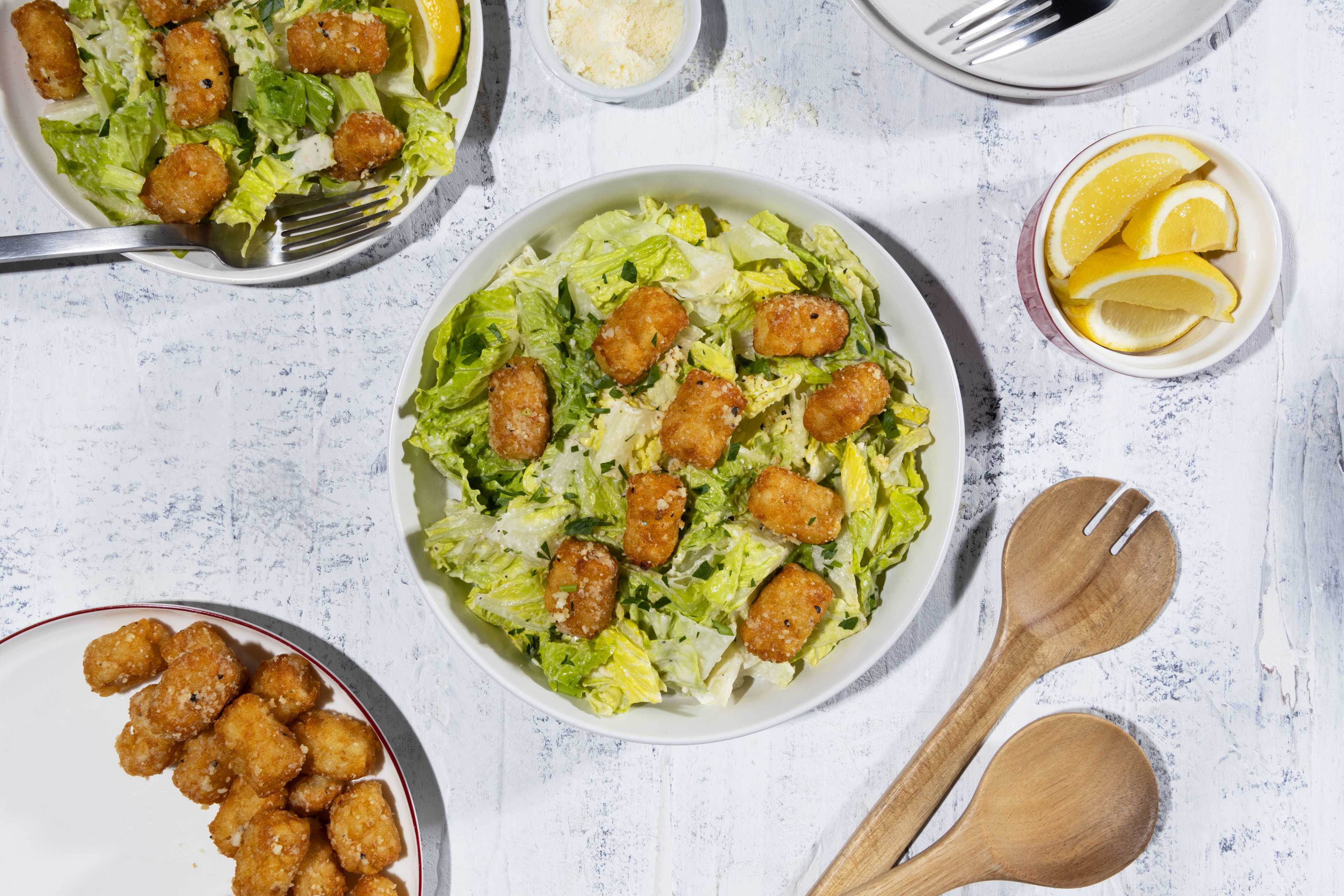 A top-down view of a delicious Caesar salad featuring crispy Ore-Ida Tater Tots. Two bowls of salad are presented on a textured white surface, alongside a plate of Tater Tots, a small bowl of grated parmesan cheese, lemon wedges, and wooden serving spoons. The salads consist of fresh, green romaine lettuce, creamy Caesar dressing, and golden-brown Tater Tots.