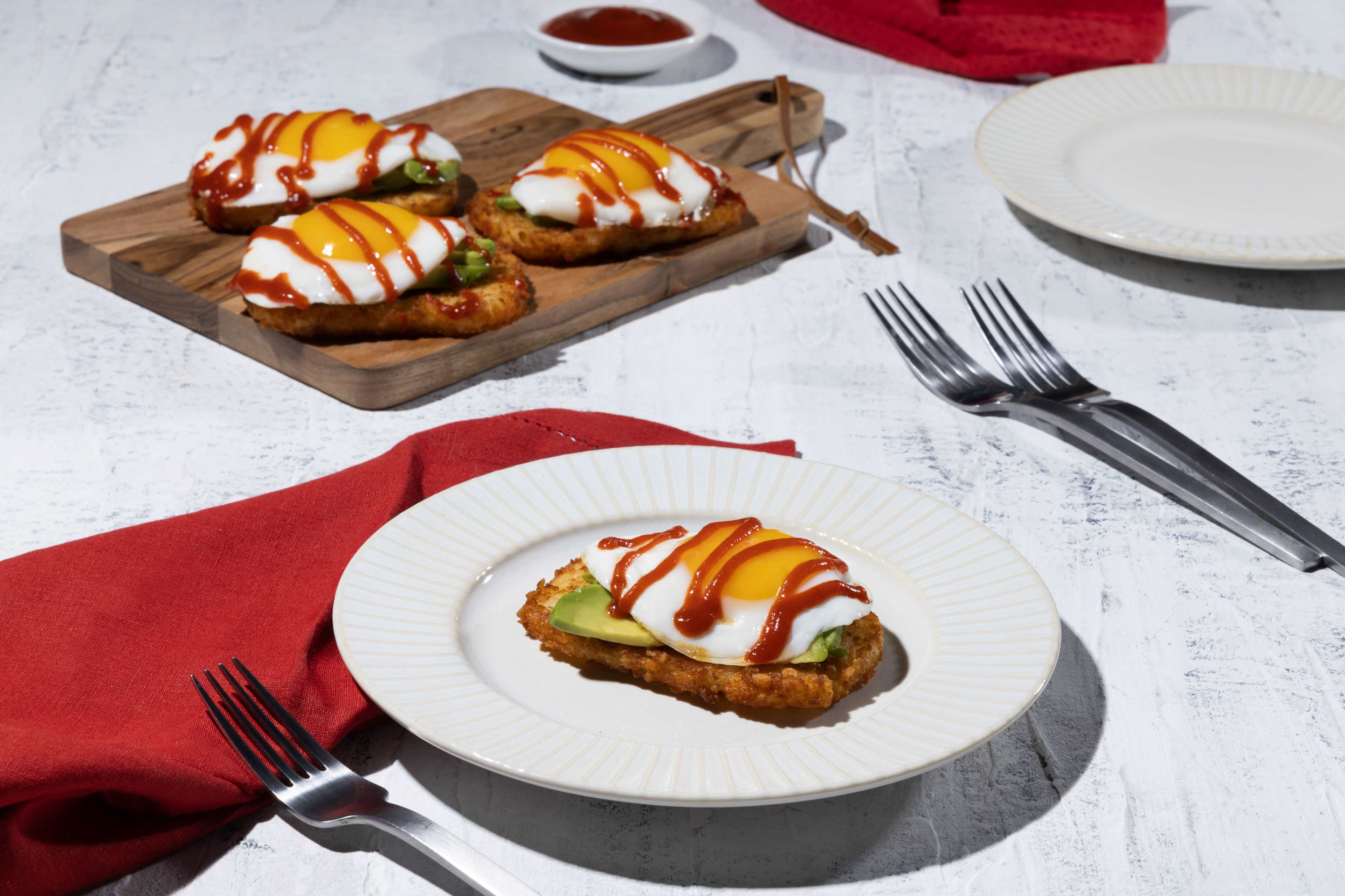 A close-up overhead shot of a delicious breakfast or brunch: hashbrown avocado toast. A crispy golden-brown hashbrown patty is topped with creamy avocado slices and a perfectly fried egg. A generous drizzle of ketchup adds a vibrant red pop of color. The main toast is presented on a textured off-white plate with ridges, accompanied by a red napkin and silverware. In the background, a wooden cutting board holds three more similarly prepared toasts, ready to be served. A small white bowl of ketchup is visible in the softly lit scene.
