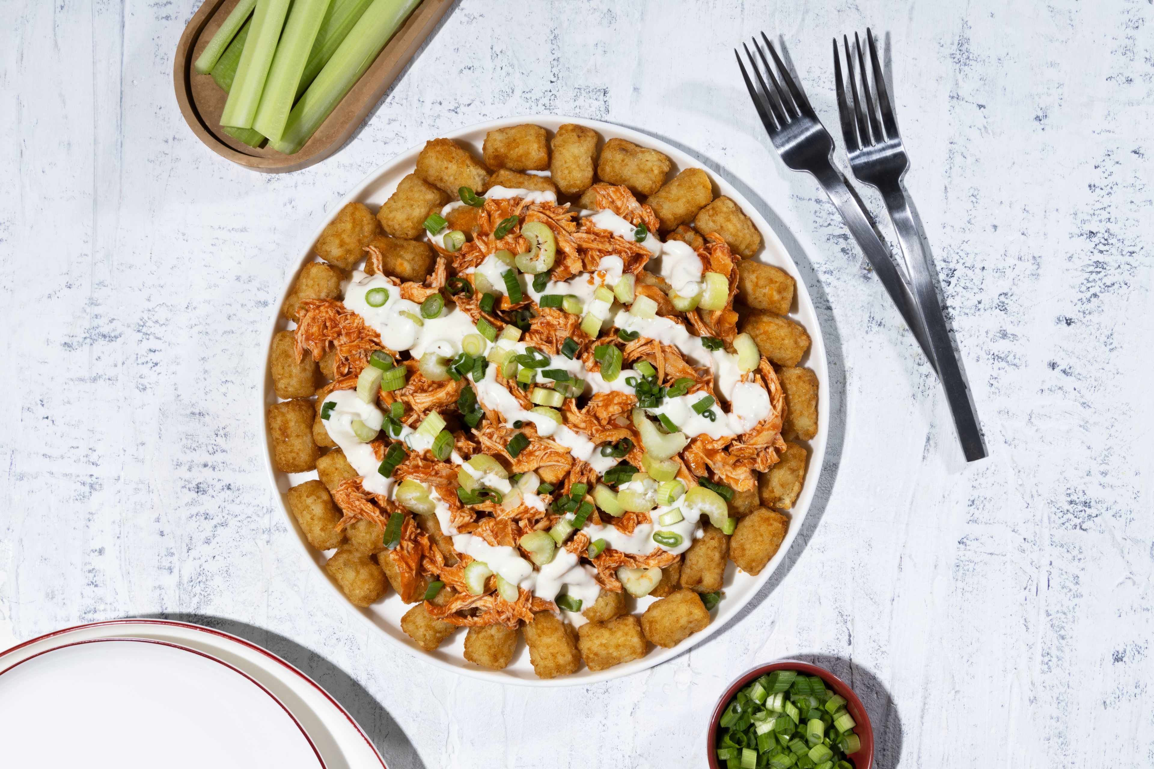 A top-down view of a plate of buffalo chicken totchos. Tater tots are arranged around the edge of the plate with shredded buffalo chicken in the center, topped with a creamy sauce, chopped celery, and green onions. Two forks are placed to the upper right, a small bowl of chopped green onions is at the lower right, and a dish of celery sticks is at the upper left, all on a textured white surface.