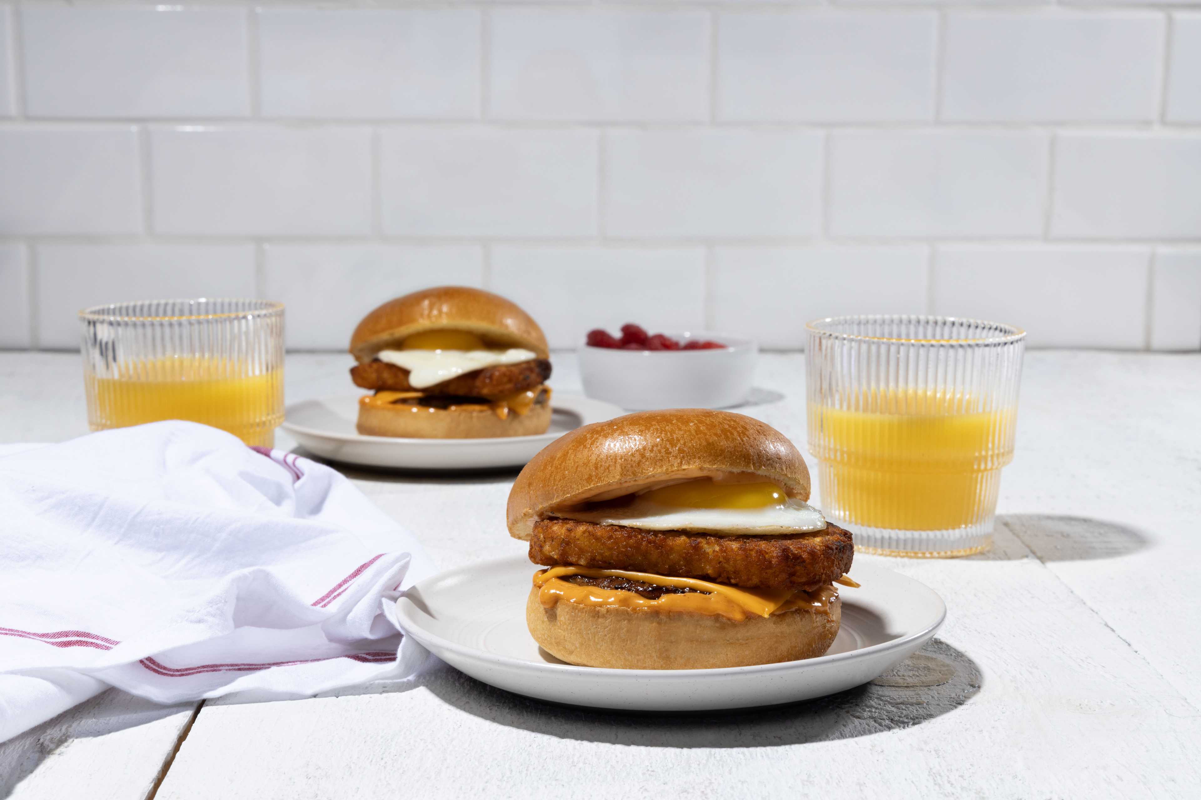 Two delicious breakfast sandwiches featuring crispy hashbrowns, a perfectly fried egg, and a creamy sauce, served on toasted brioche buns. Two glasses of orange juice and a small bowl of raspberries complete the breakfast setting on a bright white wooden table.