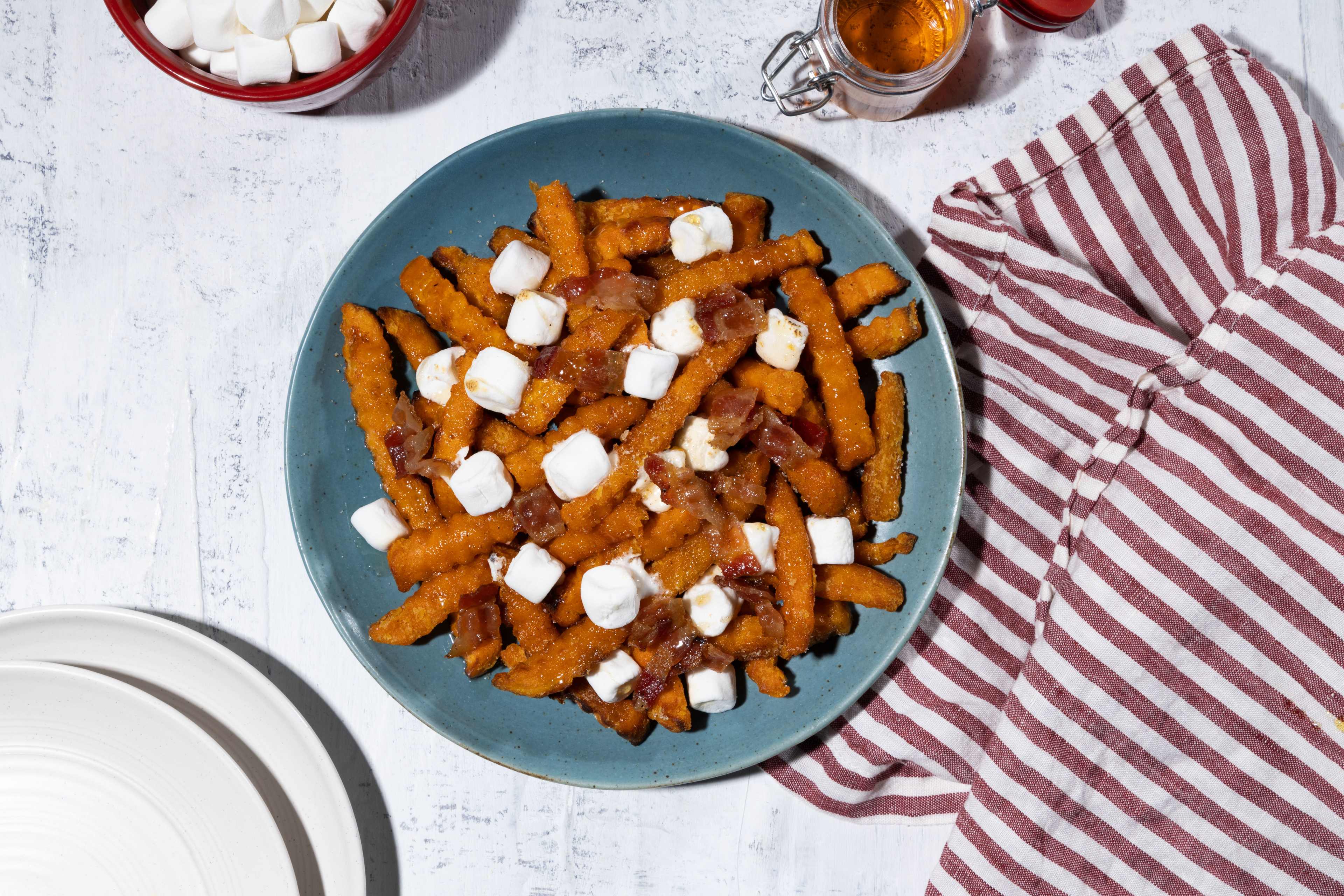 A top-down close-up view of a plate of sweet potato poutine. Crispy sweet potato fries are coated in a rich sauce and topped with fluffy marshmallows and crumbled bacon. The poutine sits on a speckled light-colored surface, with a red bowl of marshmallows, a small jar of syrup or honey, and a striped reddish-brown and white cloth napkin arranged around it. A stack of white plates is partially visible at the bottom left corner of the frame.